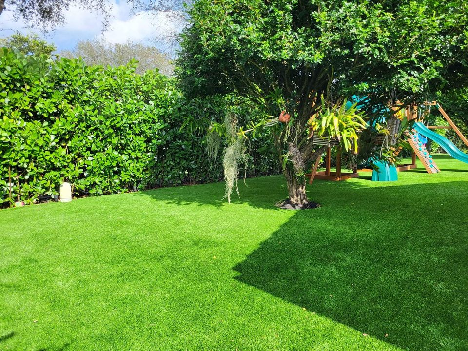 A lush green backyard in Albuquerque features vibrant artificial grass, a large tree adorned with hanging plants, and a playset with a blue and orange slide. Dense bushes form a hedge against the bright, partly cloudy sky.