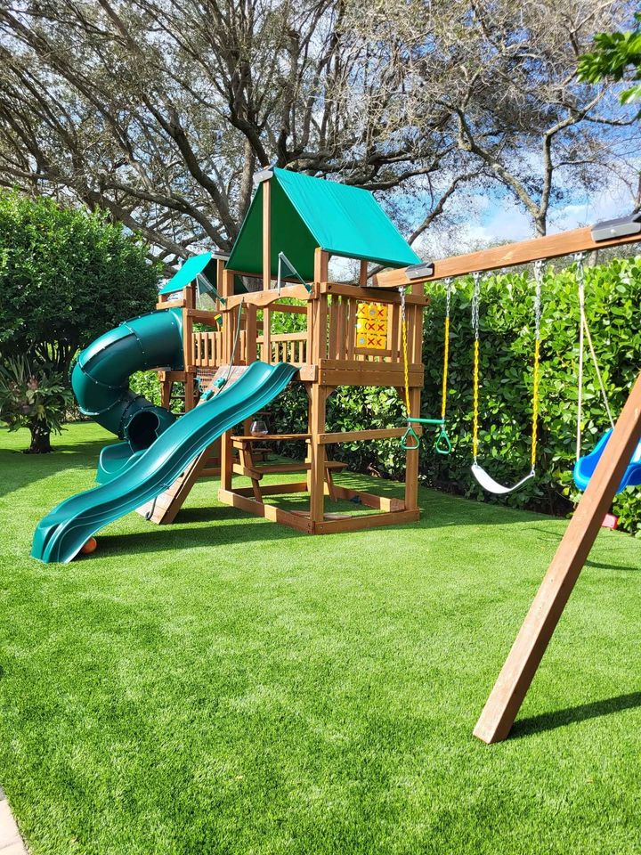 A wooden playground set with a green slide, climbing wall, and swings is nestled on lush ABQ Turf. Trees and bushes surround the area, offering a vibrant backdrop under a clear blue sky.