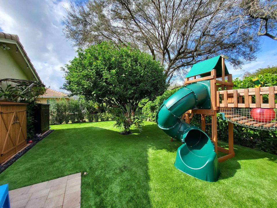 A vibrant backyard in Albuquerque with a lush green lawn and a large leafy tree complements the wooden playset, featuring a green spiral slide. The artificial grass enhances the peaceful, sunny atmosphere under the partly cloudy sky.