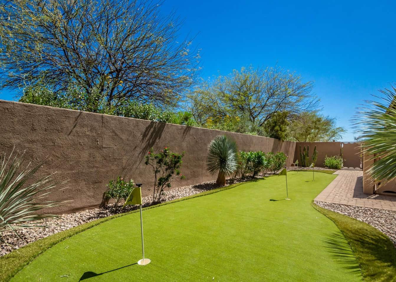 A backyard putting green featuring ABQ Turf's artificial grass installation is surrounded by desert landscaping with small plants and trees against a brown stucco wall under a clear blue sky.