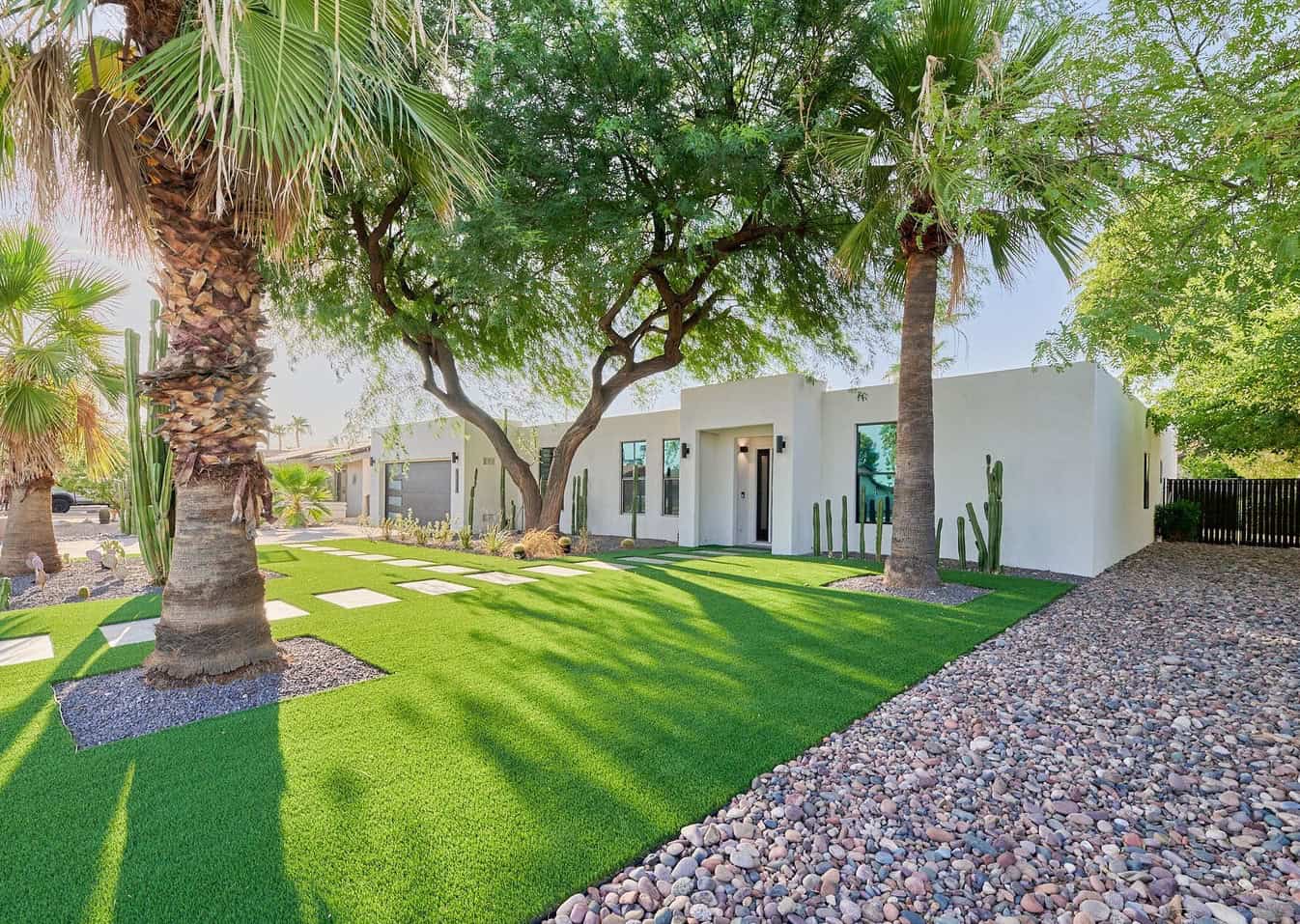 Modern white house with clean lines surrounded by tall palm trees and lush green landscaping. A pathway of stepping stones leads to the entrance, complemented by ABQ Turf's pet-friendly artificial grass, seamlessly blending with the vibrant greenery for a pristine look.