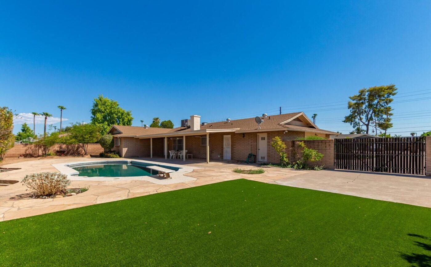 A charming backyard in Albuquerque, NM, features a kidney-shaped pool encircled by a stone patio. A cozy table sits on a covered patio attached to the single-story brick house. The yard's greenery is enhanced with lush artificial grass, and the sky remains clear and blue.