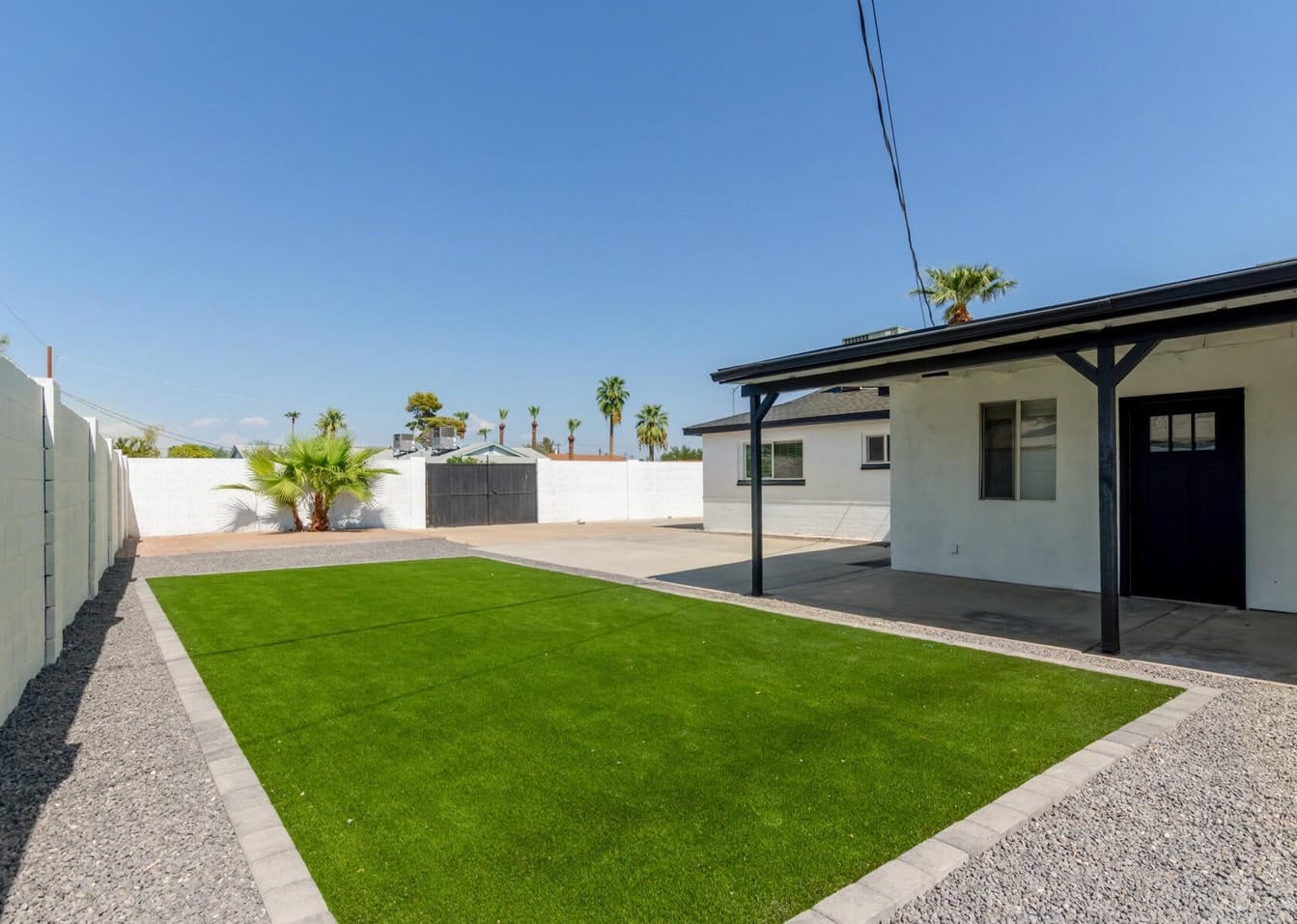 A backyard features ABQ Turf, offering a lush green lawn bordered by gravel and a concrete patio area. A small white building with a black door and pillars stands to the right. Pet-friendly artificial grass surrounds palm trees against the clear blue sky in the background.