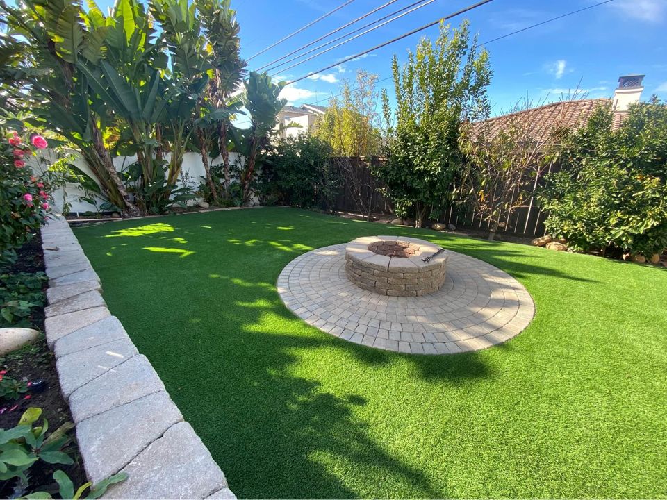 A backyard featuring ABQ Turf's lush synthetic turf, a circular stone fire pit at its heart, surrounded by mature trees and plants. A stone border lines the immaculate grass, all under a clear blue sky.