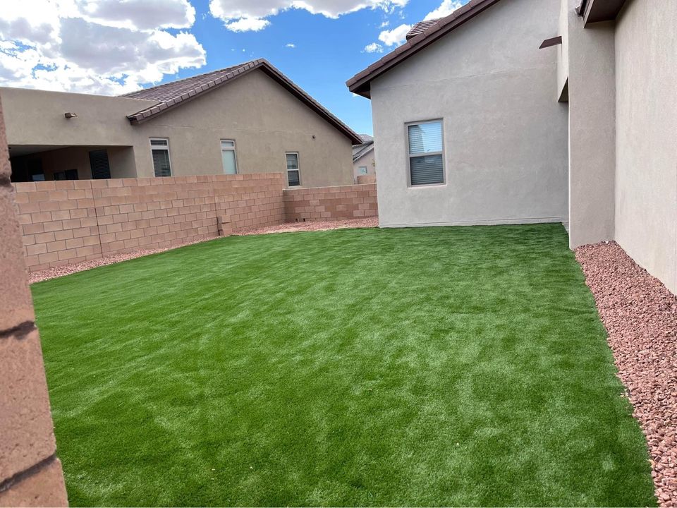 The backyard landscape features lush artificial grass from ABQ Turf, encircled by a beige stone wall and nearby houses. Brown gravel borders the green expanse, while above, the sky remains partly cloudy.