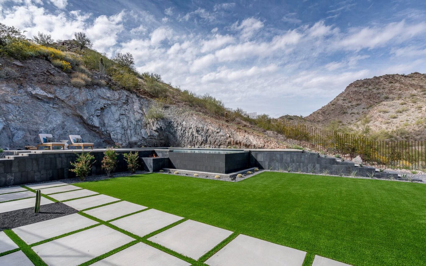 A modern backyard featuring a manicured lawn of synthetic turf with geometric concrete tiles. In the background, rocky hills rise under a partly cloudy sky, complemented by two lounge chairs on a patio. Sparse desert vegetation lines the hills, adding to the tranquil ABQ Turf setting.