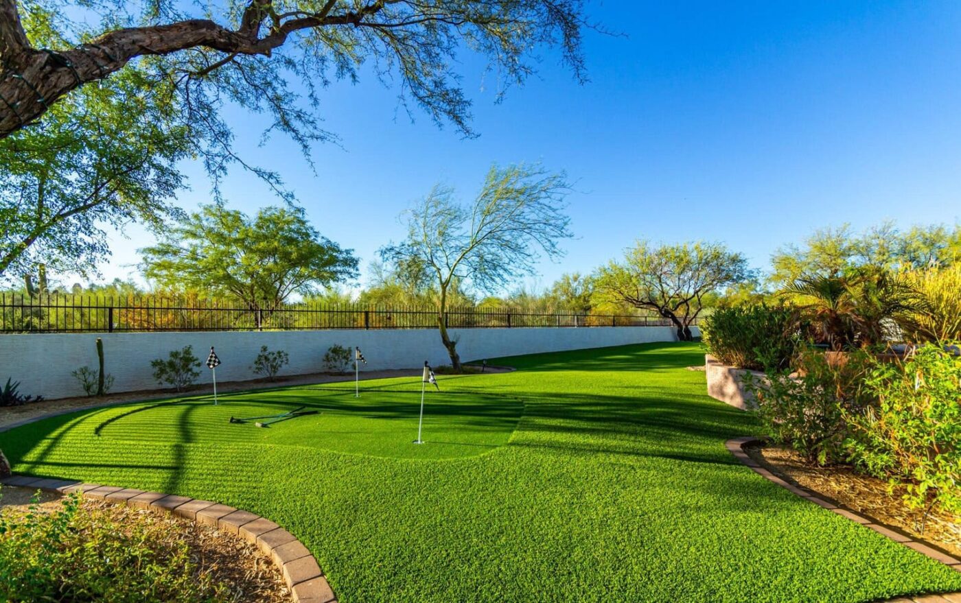 A scenic backyard in Albuquerque, NM, features a mini-golf course with three flags, surrounded by lush artificial grass and bordered by trees and shrubs. The clear blue sky enhances the vibrant greenery of the landscape.