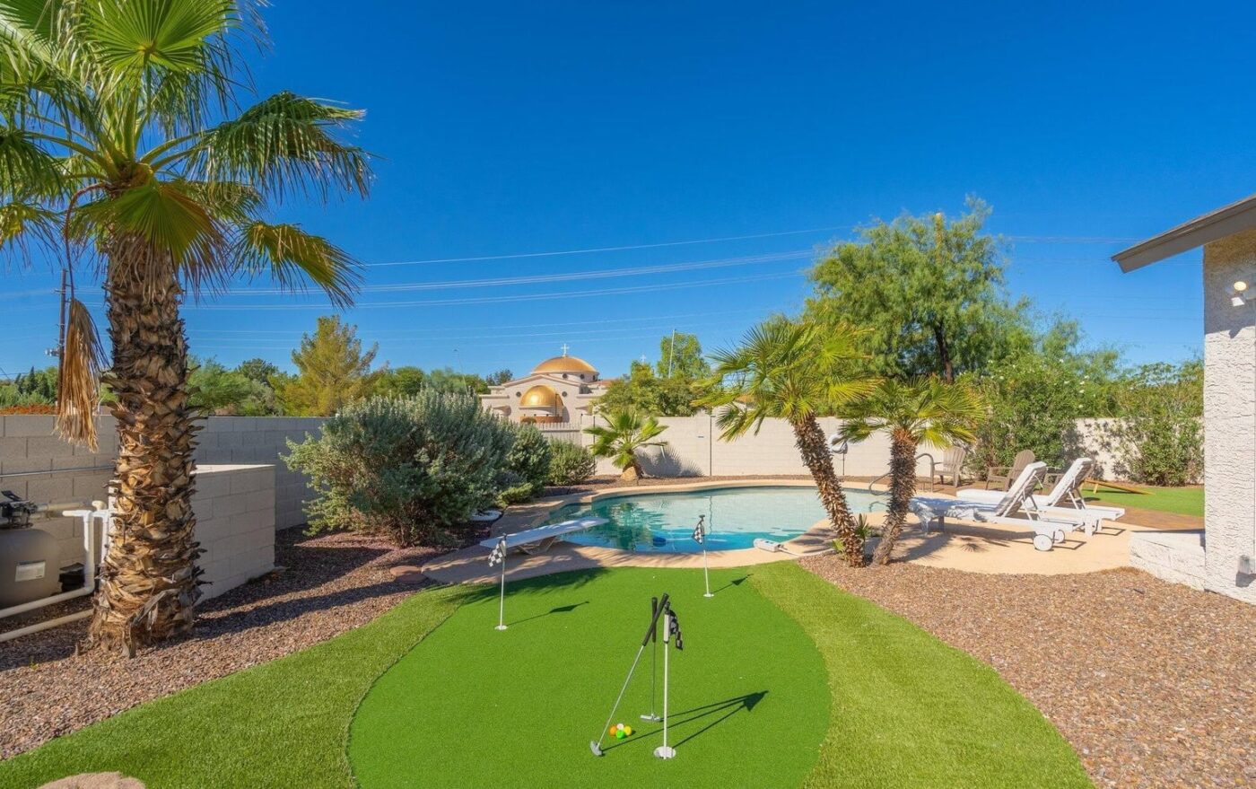 A backyard scene showcases a small putting green with golf clubs and palm trees, highlighting the expertise of ABQ Turf in artificial grass installation. In the background, a round swimming pool with lounge chairs sits under a clear blue sky, completing this oasis.