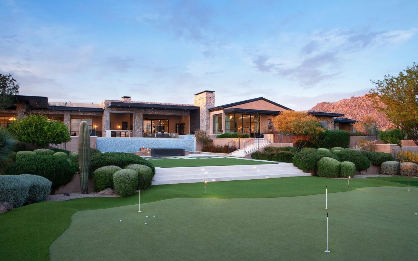 A luxurious modern house in Albuquerque features large glass windows and stone accents, surrounded by landscaped greenery and desert plants. In the foreground, a well-maintained putting green with pet-friendly artificial grass is visible. Mountains lie in the distance under a partly cloudy sky.