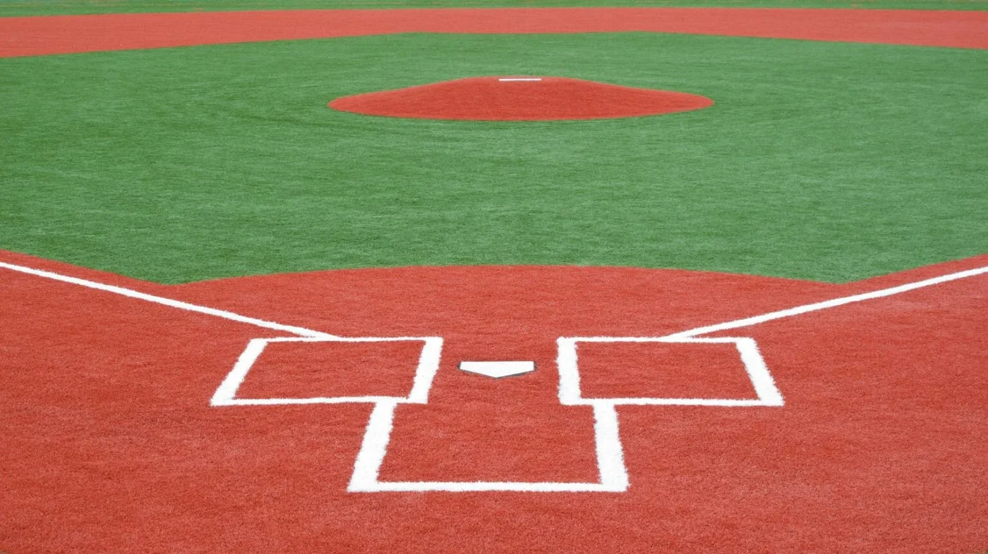 A baseball field featuring a red clay pitcher's mound and home plate area, complemented by white markings on the artificial sports turf. The image captures the geometric layout of the field, focusing on the home base and pitcher's mound.