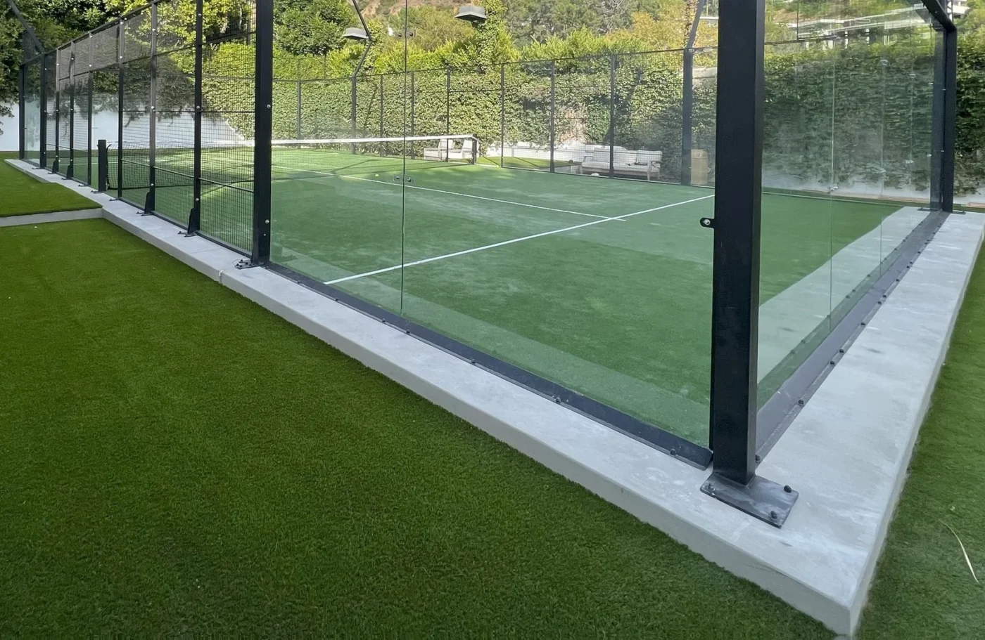 A glass-enclosed paddle tennis court boasts bright green artificial sports turf, resembling putting greens. The area is adorned with trimmed grass, surrounded by trees and hedges under Albuquerque NM's clear sky.