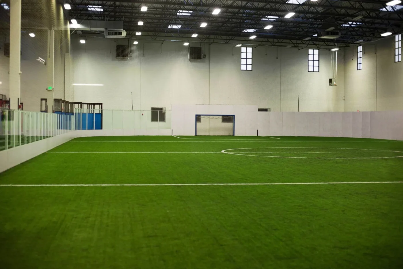 An indoor soccer field with artificial sports turf, surrounded by white walls and glass panels. High ceiling with visible beams and ceiling lights, and a goalpost at the far end. The empty and well-lit space offers an ideal environment for players to excel in their game.