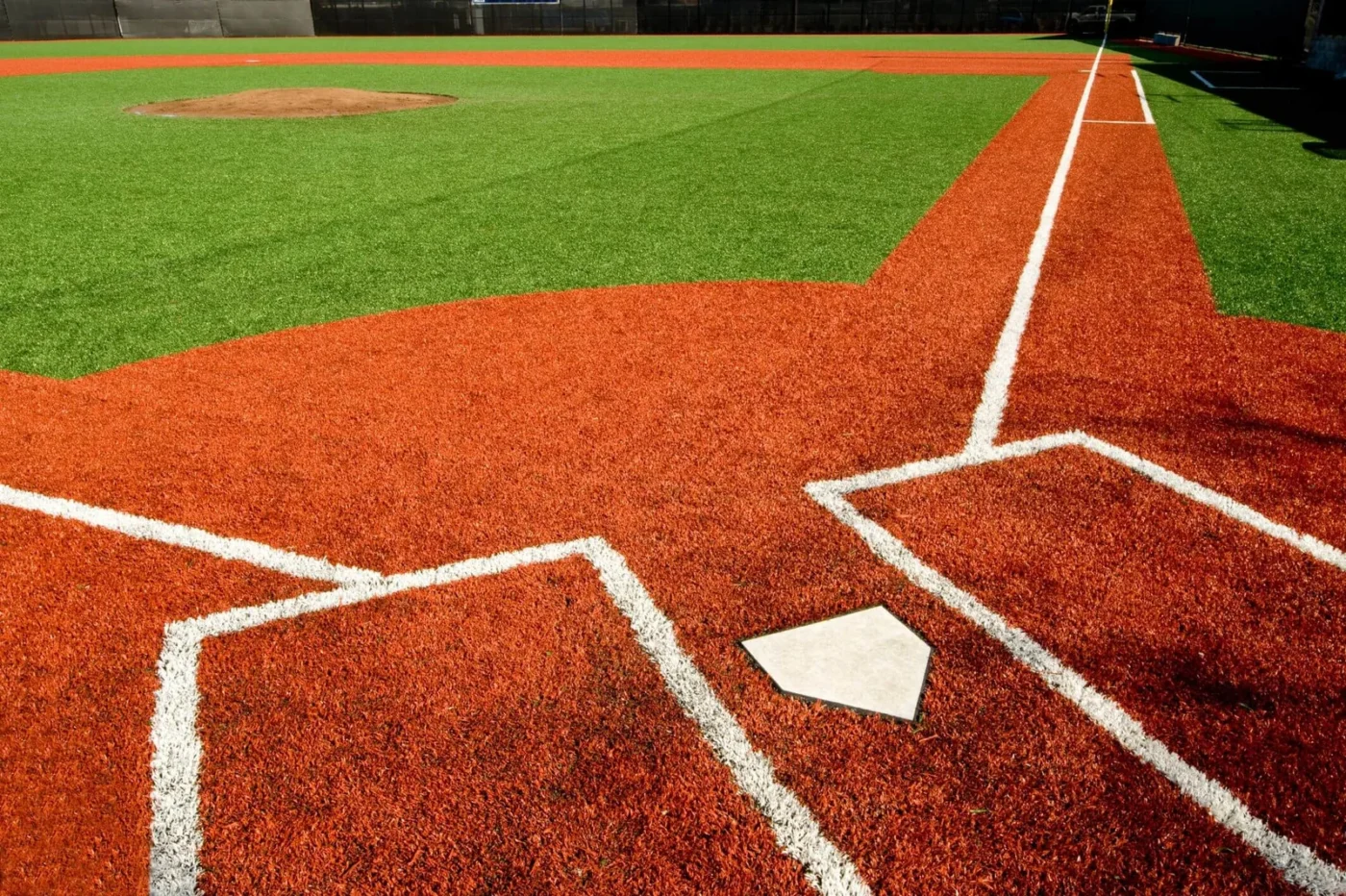 A baseball field in Albuquerque showcases a home plate and pitcher's mound on bright green artificial sports turf, surrounded by red turf batter's boxes and baseline. White lines mark the edges, contrasting with the colorful surface. Sunlight casts shadows reminiscent of putting greens on the field.